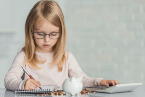 Little girl wearing eyeglasses writes in notebook at table witj calculator , coins and piggy bank