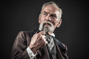 Pipe smoking vintage characteristic senior man with gray hair and beard. Studio shot against dark background.